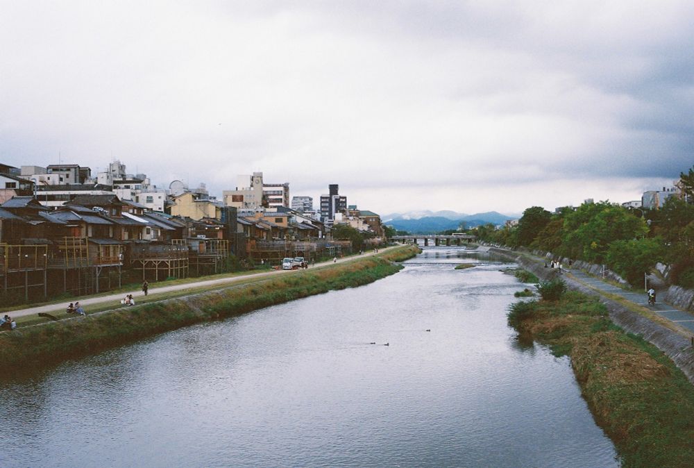 Cloudy day in Kyoto, a view on Kamo River (?) with mountain tops in the background. Some people sitting and chilling near the bank.