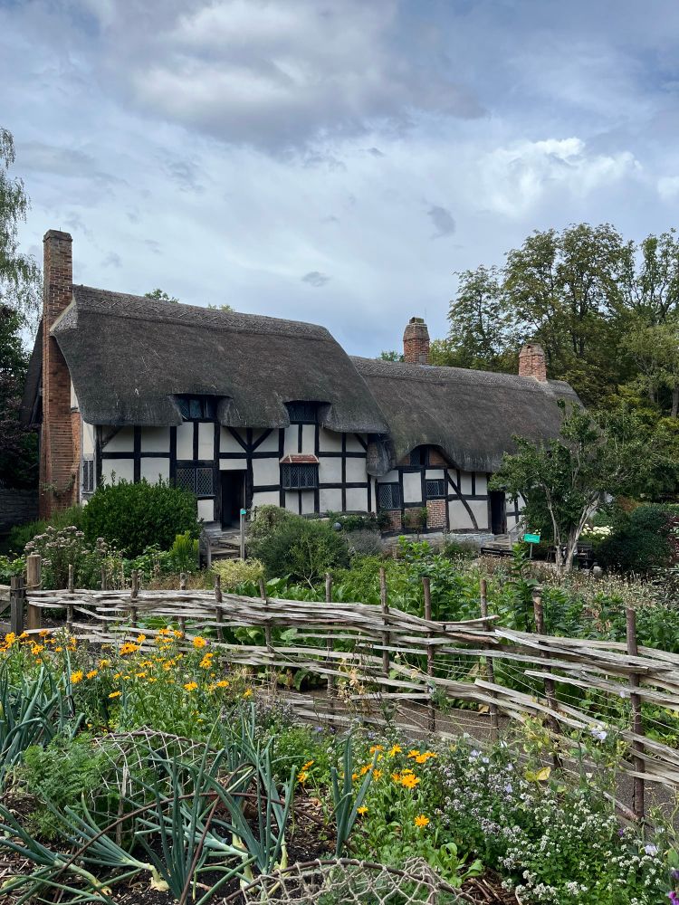 A white Tudor-styled cottage with a thatched roof and brick chimneys sits behind an extensive garden.