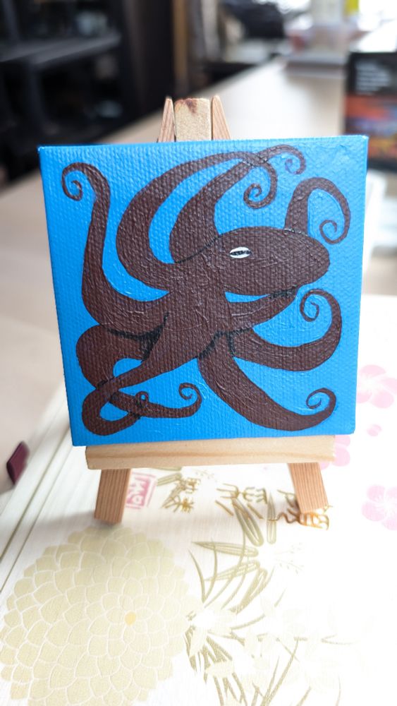 A miniature canvas and easel. The canvas has a painting of a dark red octopus on a bright blue background 