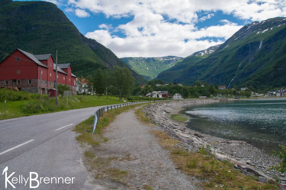A road curving around a fjord heading towards the mountains with a red building on the side