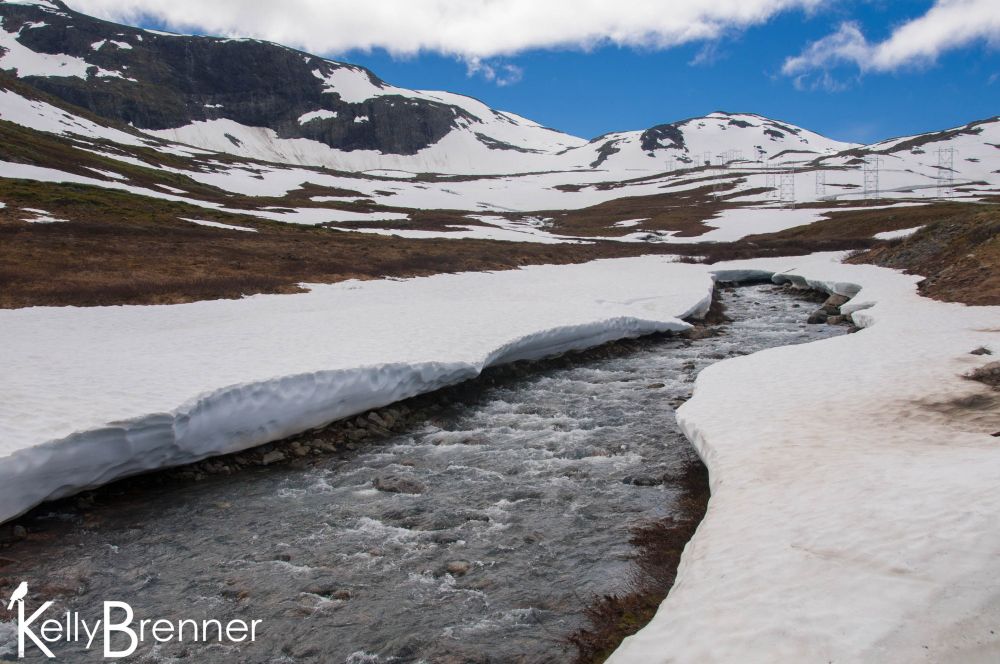 A small stream running through a snow bank with low  snow-covered hills