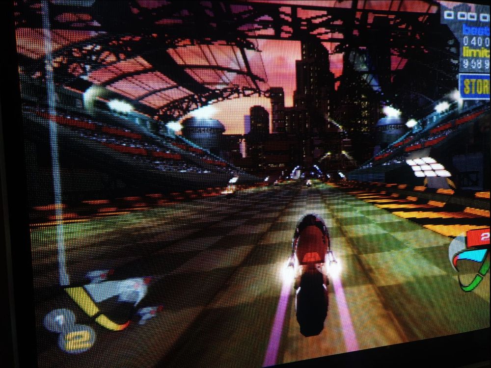 A CRT TV screenie of a third-person cyberpunk racing game with a sci-fi metropolis beneath a red sky and with a chequered section of track ahead and spotlight gantries on either side.