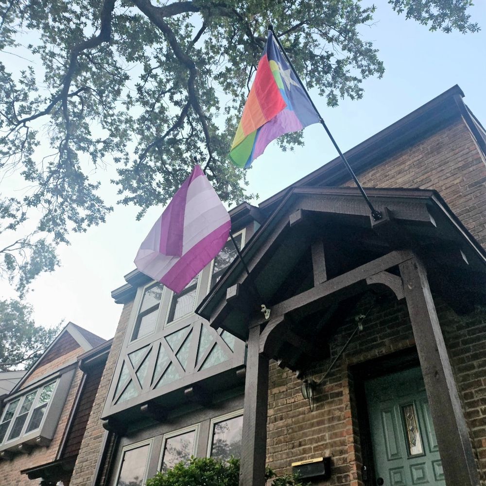 Two flags hang proudly from the front porch of a house. One is the Gray-Asexual Pride Flag, composed of five horizontal stripes: purple, gray, white, gray, and purple. This flag, created by Milith Rusignuolo in 2013, represents the nuanced space between asexuality and allosexuality—where gray symbolizes the infrequent or conditional experience of sexual attraction, white stands for allosexuality, and purple honors the asexual community. The second flag is the PRIDE Texas flag, a reimagining of the Lone Star flag where the red and white stripes are replaced with the six-color rainbow of LGBTQ+ pride. Together, these flags express a deep kavod habriyot—respect for the dignity of every person—and reflect a home that embraces both queer identity and regional pride with integrity and joy.
