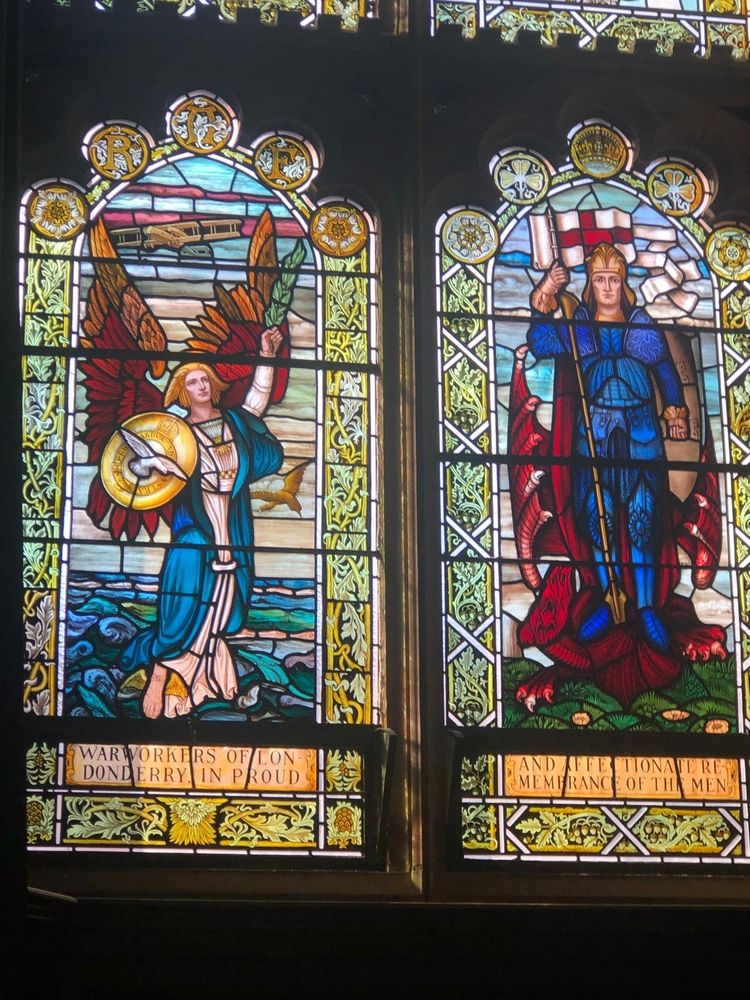 2 of the stained glass windows in the Derry Guildhall. The left features a winged figure with a blue robe & autumnal coloured wings. The right hand side one is an image of St George, in a blue (blacked) suit of armour, standing on the vanquished dragon, leaning on the pole of his pennant, which is fluttering above.