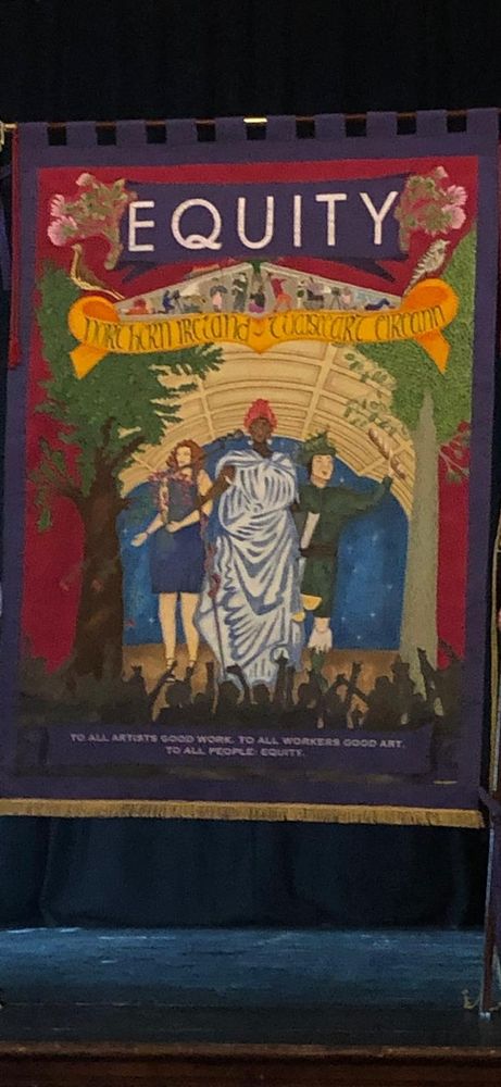 Fabric banner features a purple border with an inner rectangle of a fuschia-ish tone. At the top a purple piece, shaped like a header ribbon carries print in white: "Equity".
Beneath, 3 figures on the stage of a large hall performing to a silhouetted crowd in the bottom of the banner. To either side of the stage there are trees. One has a dark trunk, the other has a light trunk.