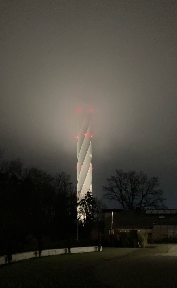 Ein angeleuchteter Turm verschwindet in den Wolken. Der Turm sieht aus als wäre er in sich gedreht. Die Wolken werden vom Licht des Turms erleuchtet.