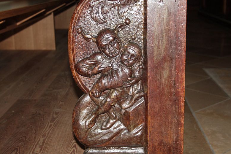 Bild im Querformat: In der Bildmitte ist die hölzerne Stuhlwange einer Kirchenbank zu sehen. Eine geschnitzte Relieff-Figur mit Narrenkappe hält ein altmodisch gewickeltes Kind im Arm, das ebenfalls eine Narrenkappe mit Glöckchen trägt. Die große Figur, die Narrenmutter, führt einen Löffel zum Mund des Kindes.