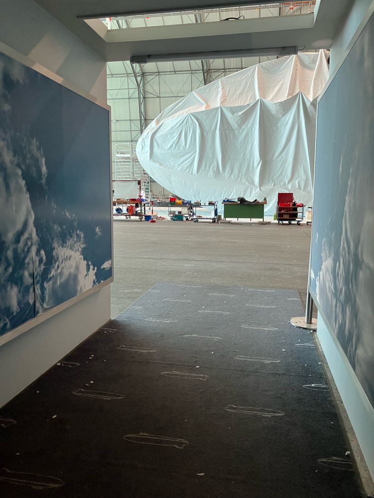 Blick durch einen Gang in eine Werkshalle. Weiter hinten in der Halle sieht man den vorderen Teil eines Zeppelins, dessen Hülle noch sehr locker und faltig liegt. Davor stehen ein paar bunte Kisten und Rollkästen, vermutlich mit Werkzeug.