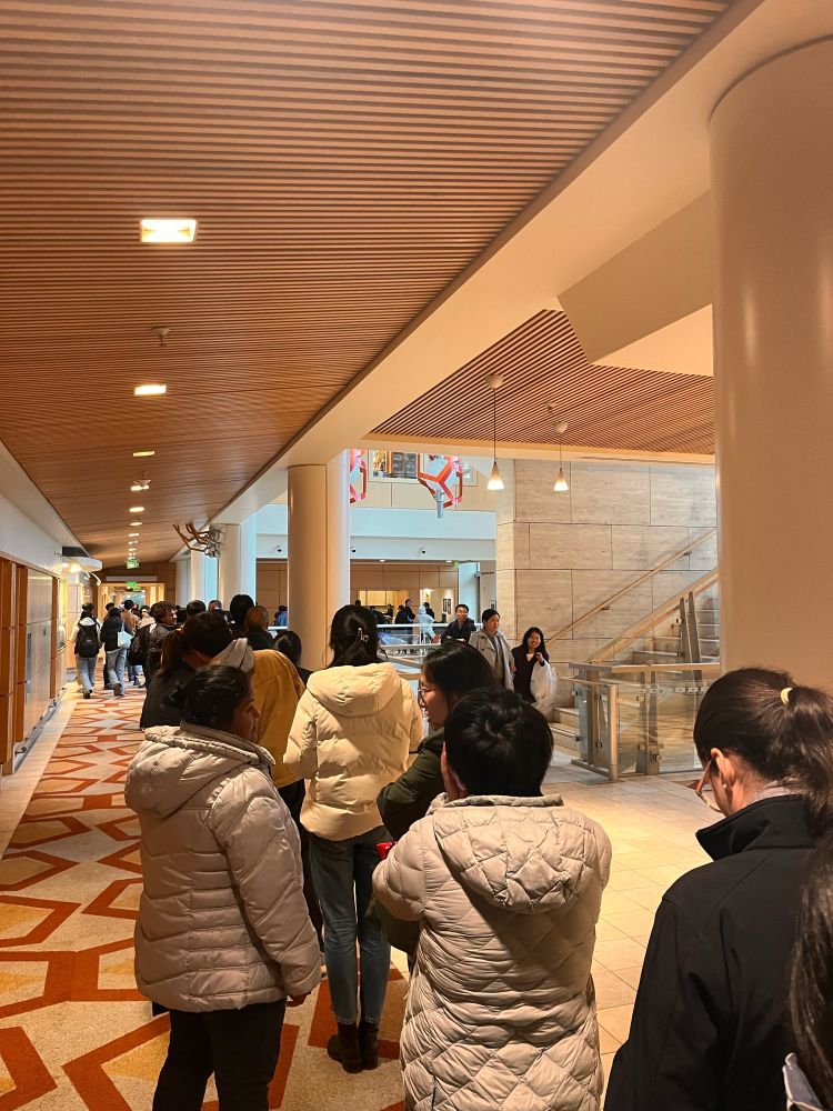 A crowd of people wraps around a hallway. About 25-30 people are visible