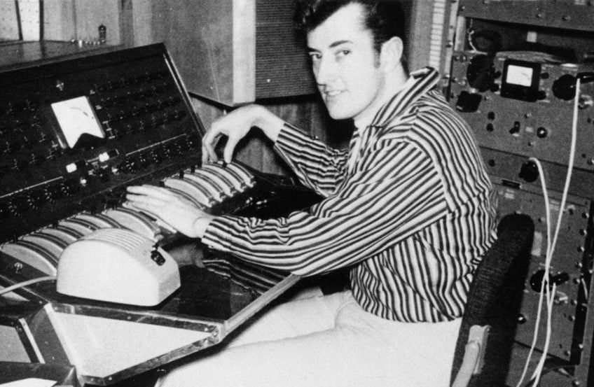 Black and white photograph of Joe Meek, sitting at what I assume to be an old audio pult, with his hands on the faders. He wears a stripped button down and light trousers and is turning his head towards the camera with a slight smile