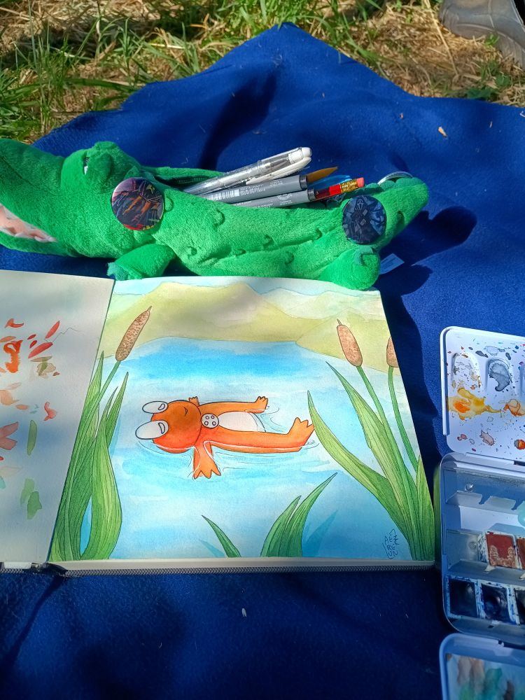 A picture of a sketchbook with a crocodile pencilcase and a watercolor box next to it, on a blue plaid on the grass. The sketchbook has a painting of a little cartoon orange frog laying in the water of a montain lake with reeds in front.