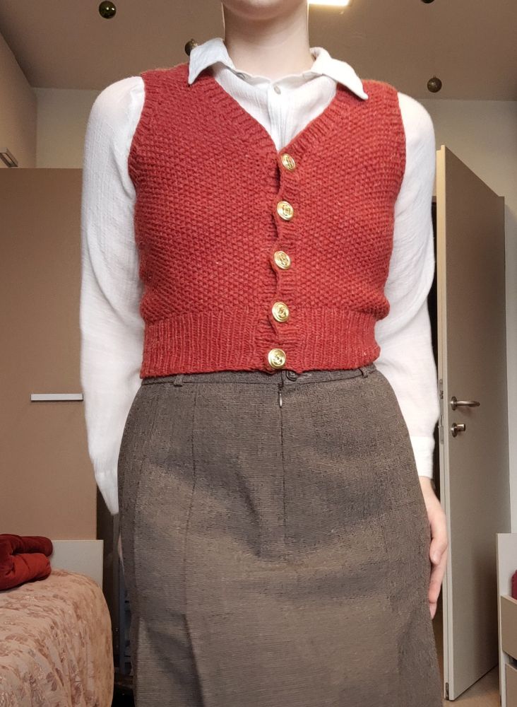 Person in frame from chin to thighs. Dressed in a light shirt, brown wool skirt and a handknit red button up vest. Awkwardly posing.