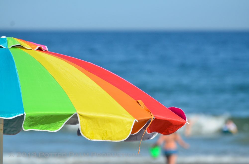 A brightly colored beach umbrella. In the background is a wave breaking and people playing in the water. 