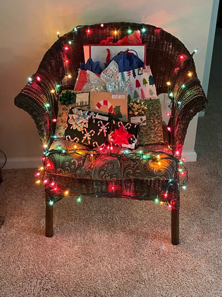 The Christmas Chair: a brown wicker chair with colored mini lights wrapped all around the frame and wrapped gifts arrayed on the seat cushion.