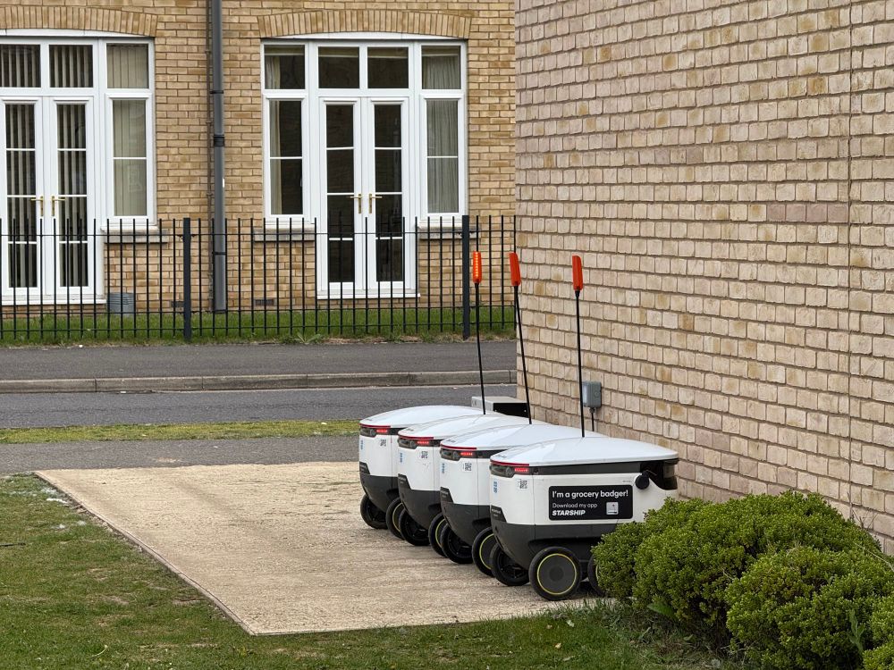 A photograph of food delivery ‘starship’ robots reverse parked in a neat line along a sand coloured brick wall in a residential area. 
