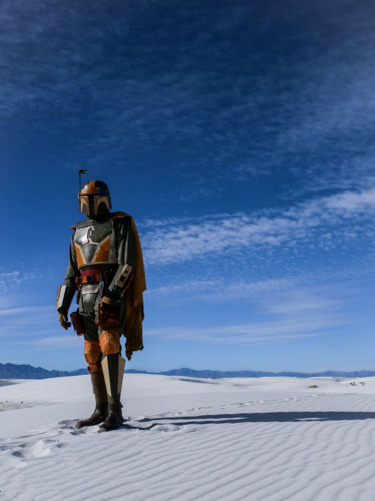 A Mandalorian in green, terracotta, and beige standing against the white sands and blue skies of White Sands National Park in New Mexico. 