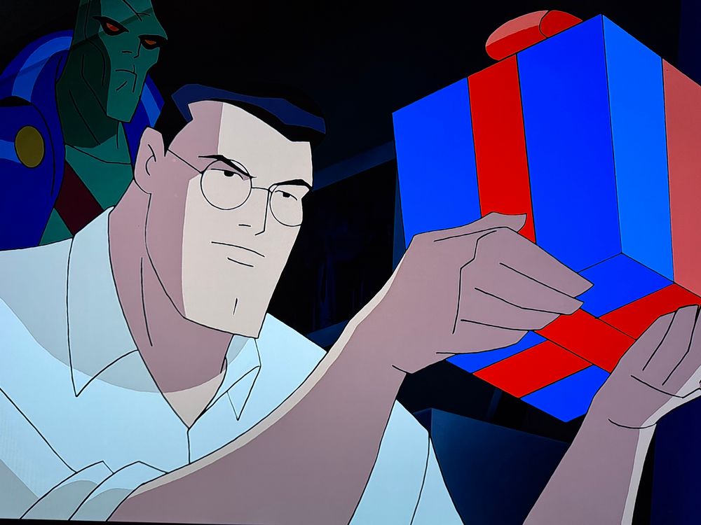 Picture of a TV screen. A still from the justice league animated holiday special. Clark Kent is staring at a present, while a ghostly invisible Martian manhunter observes behind him, unnoticed.