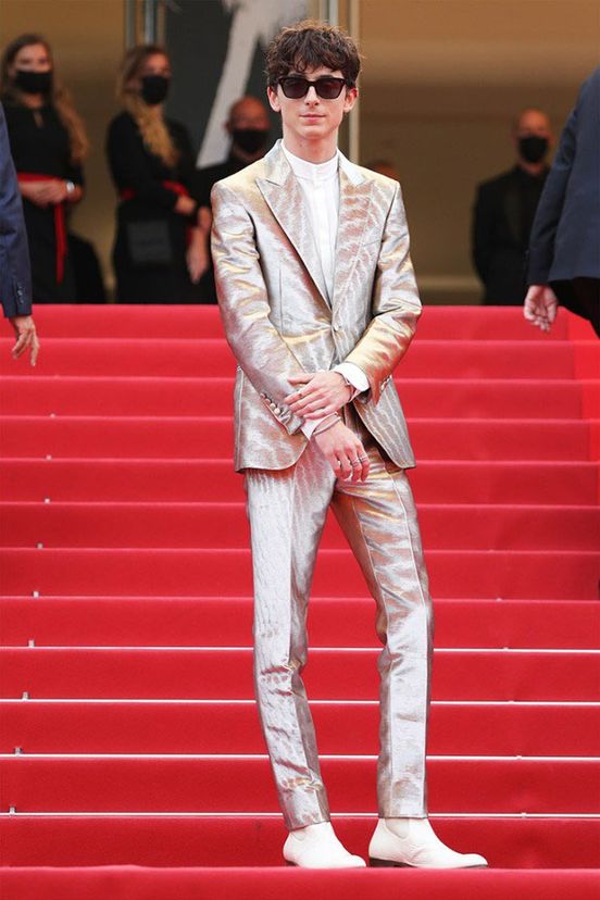 Timothée Chalamet at Cannes 2021 standing on a red staircase wearing sunglasses, a white crewneck shirt, a shiny metallic suit, and white boots. Behind him are out of focus staff wearing black Covid masks.