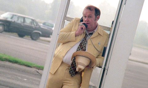 Actor M. Emmet Walsh in the film Blood Simple, looking heavy and sweaty in a phone booth, wearing a yellow suit and holding a cowboy hat.