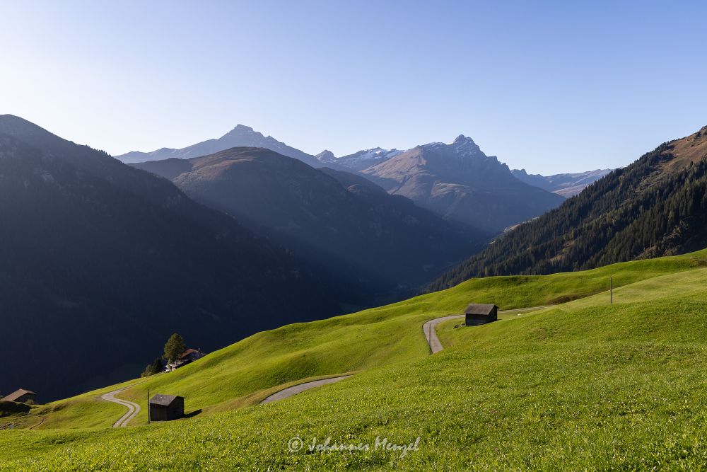 Morning light on an alpine landscape, looking down into a valley. Green pasture in the foreground, with a narrow winding road and a couple of barns, and high mountains in the background.