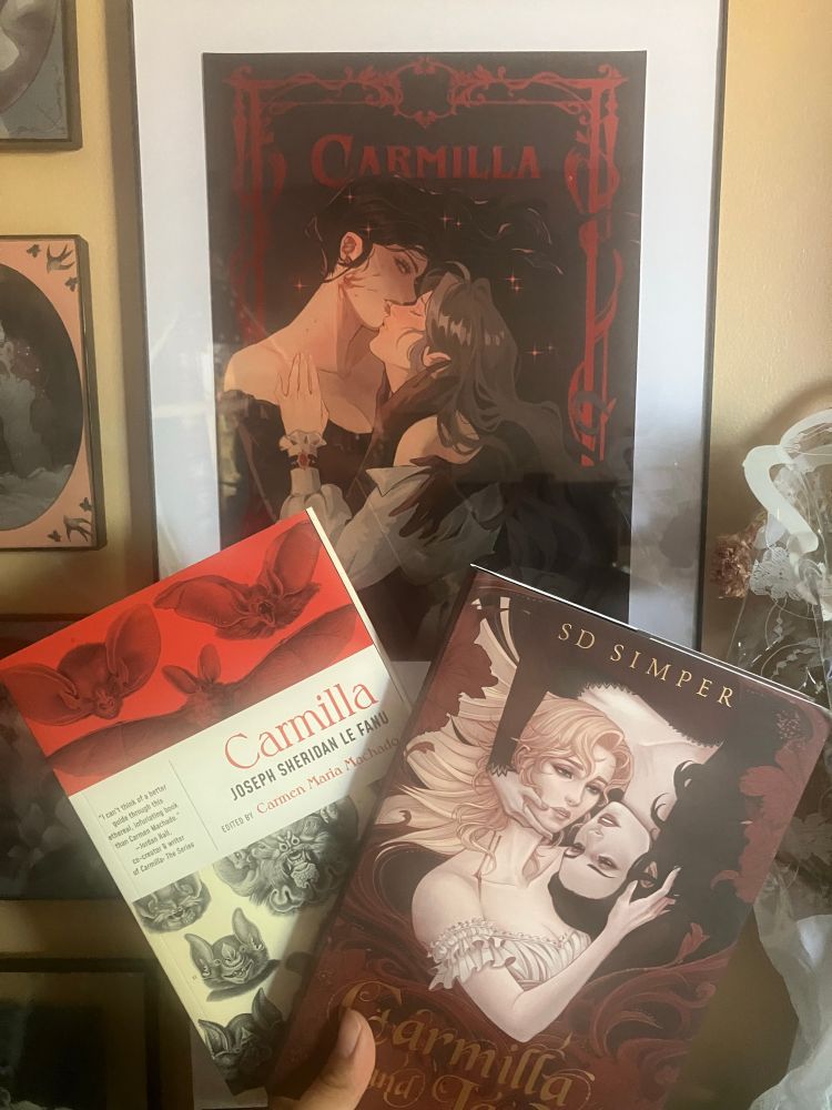 Photo of a framed print of Camilla and Laura kissing, with two books being held in front: Carmilla edited by Carmen Maria Machado, and Carmilla and Laura by SD Simper