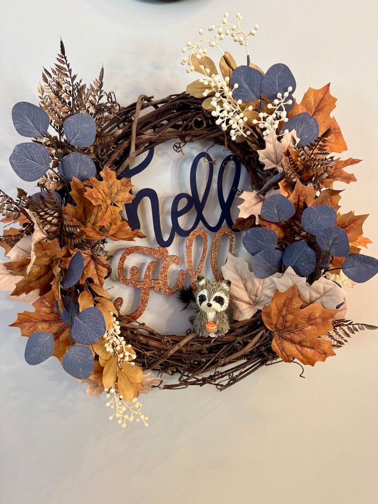 Grapevine wreath with brown and cream leaves, cream berries, blue eucalyptus, and brown ferns. Letters spell hello fall and a small raccoon with a pie sits at the bottom. 