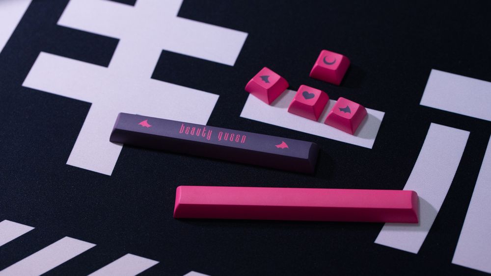 some keycaps from my custom keycap set: a dark purple spacebar with the text "beauty queen" between two little demon wings is surrounded by hot pink keycaps with dark purple legends