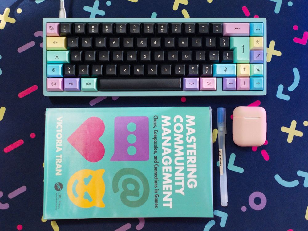 on a colorful deskmat, a colorful mechanical keyboard, a muji pen, my airpods, and Victoria Tran's community management book (all in matching color palettes!)