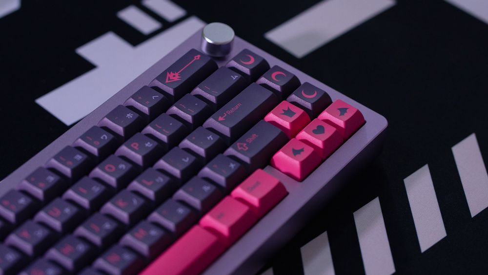 a set of custom dark purple keycaps with hot pink legends and accents on a purple 65% keyboard. the legends include little moons, a heart, little bat/demon wings, a crown, Etna's spear