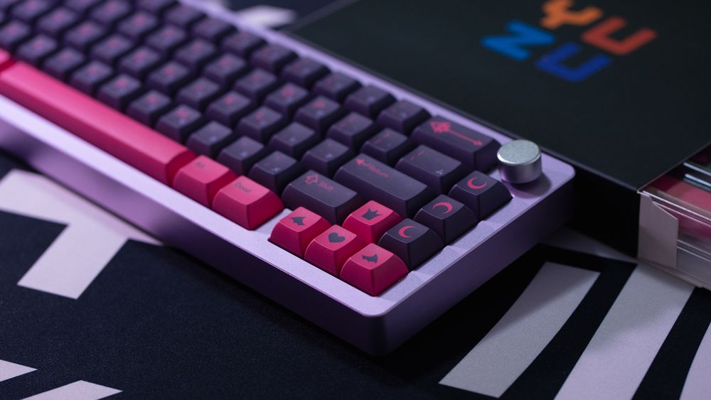 a set of custom dark purple keycaps with hot pink legends and accents on a purple 65% keyboard. behind the keyboard, the YUZU keycaps box the set came in