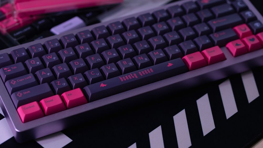 a set of custom dark purple keycaps with hot pink legends and accents on a purple 65% keyboard. "beauty queen" is written on the spacebar, between two little bat/demon wings
