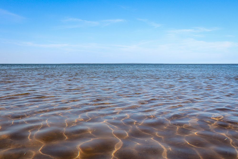 Calm, shallow ocean with a sandy, orange bottom. Blue sky with wispy clouds above. Pictou Island barely visible in the extreme distance.