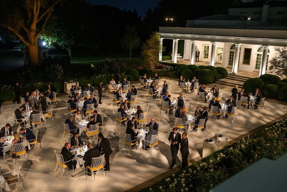 The area of the White House previously known as the Rose Garden. This area is now covered with cement and has a dozen cafe tables arranged in diagonal lines. People sit at the tables, drinking and eating at night and the area has been lit.