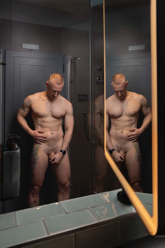 A naked muscular man is looking down at his physique with one hand covering his cock. His reflection is shown in the mirror beside. 