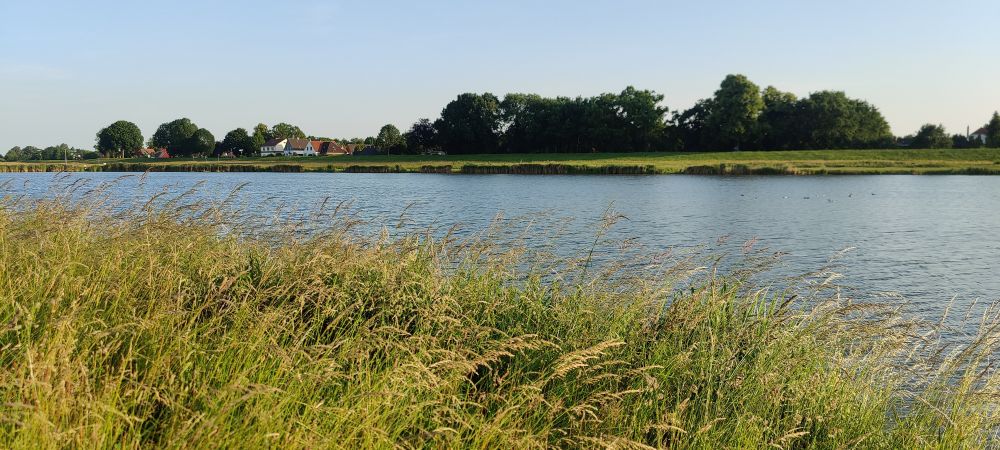 Panoramic photo of a river with grass in foreground and small houses in the background.