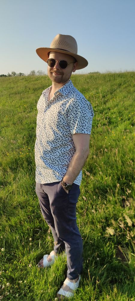 Picture of me wearing a white shirt with squiggly black pattern, dress pants, white sandals, straw Panama hat and round sunglasses. I'm standing on a grassy dike.