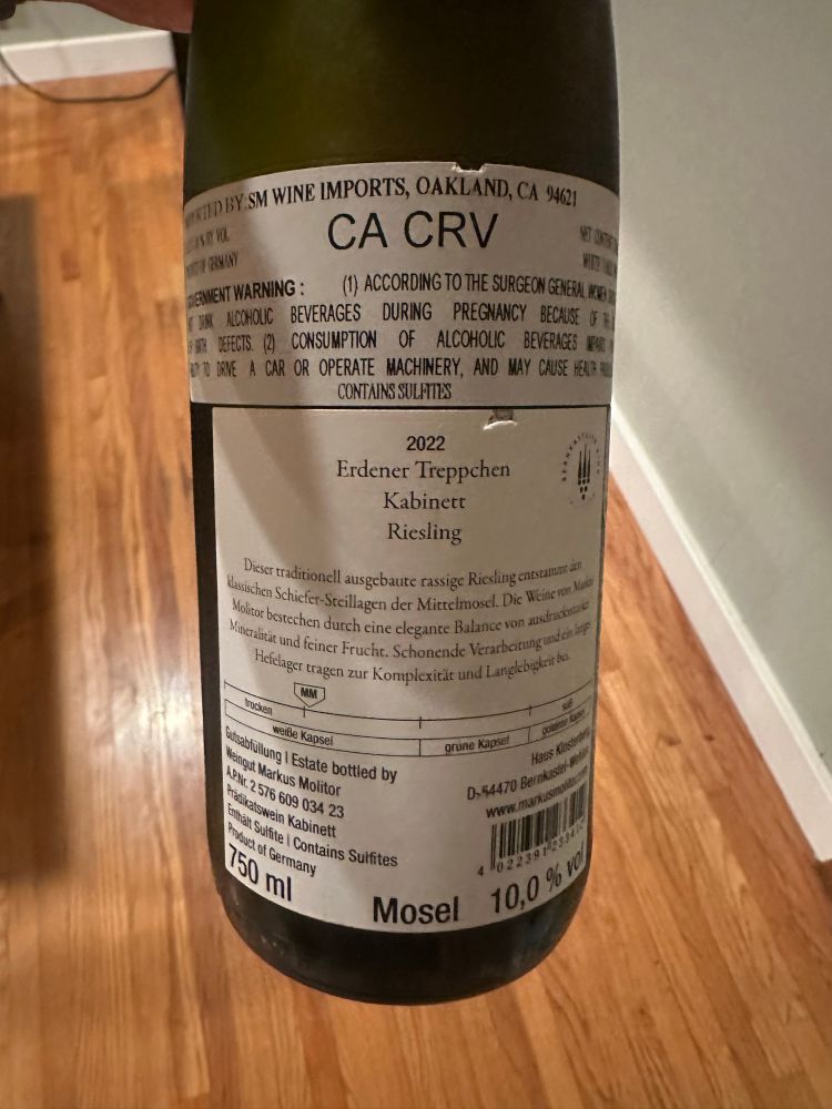 The rear label of the wine bottle.

Scanned from label:

“KENDBY SM WINE IMPORTS, OAKLAND, CA 4621
CA CRV
BENENT WARNING :
(1) ACCORDING TO THE SURGEON GENERAL NE
T SA ACCHOLC BEVERAGES DURING PREGNANCY BECAUSE
DEFECTS. (2)
CONSUMPTION OF ALCOHOLIC BEVERAGES ERS
PTO DIVE A CAR OR OPERATE MACHINERY, AND MAY CAUSE HEAD E CONTAINS SULPTITS
2022
Erdener Treppchen
Kabinett
Riesling
Dieser traditionell ausgebaute rassige Riesing cupscatmdn Ksschen Schiefer-Steillagen der Mittelmosel. Die Weine non Nao Meltor berechen durch eine clegante Balance von aushasts Mineralitär und feiner Frucht. Schonende Verarbeitung und en bap Hefelager tragen zur Komplexität und Langlebigkeira
trocken
weiße Kapsel
Stabfillung I Estate bottled by Weingut Markus Molitor
grune Kapsel
gober de
haus Alectets
APN: 2576 609 034 23
D, 54470 Berkaste Pe
Fikatswein Kabinett cathalt Sulfte | Contains Sulfites
duet of Germany
160 m
4 02239725571
Mosel 10,0 % n”