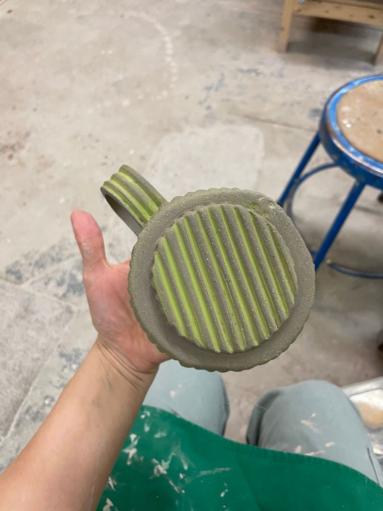 A hand holding a leather hard hand built clay mug and showing off the corrugated foot ring on the bottom, which has a chartreuse underglaze applied then wiped off the high points.
