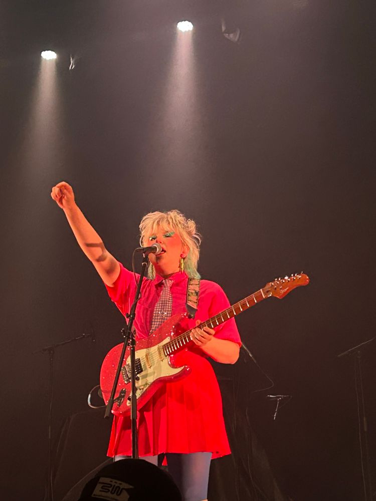 Singer on stage in all pink skirt and dress shirt with a checkered tie and blonde hair styled in a mullet. She is also holding a pink guitar and lifts her hand as she sings 