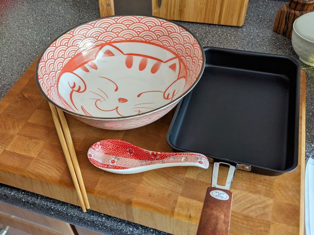 A red&white one liter noodle bowl with cute kitty in the center of it, to the right a tamagoyaki pan, at the bottom a red&white ceramic spoon that doesn't quite match the design of the bowl, but is similar enough.