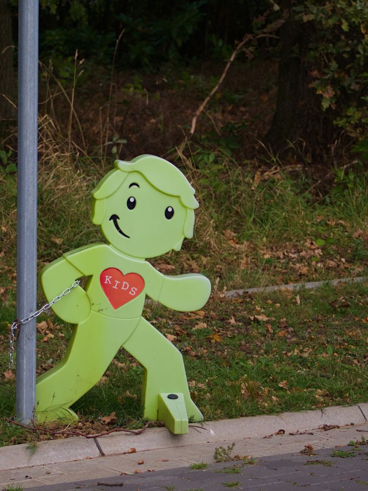 Photo of a green waist-high smirking stylized plastic child figurine with a heart in the middle of its chest reading "KIDS". The left half of the figurine is visibly discolored by prolonged sun exposure and it is chained to the post of a street sign via its arm using a steel chain and a bronze lock.