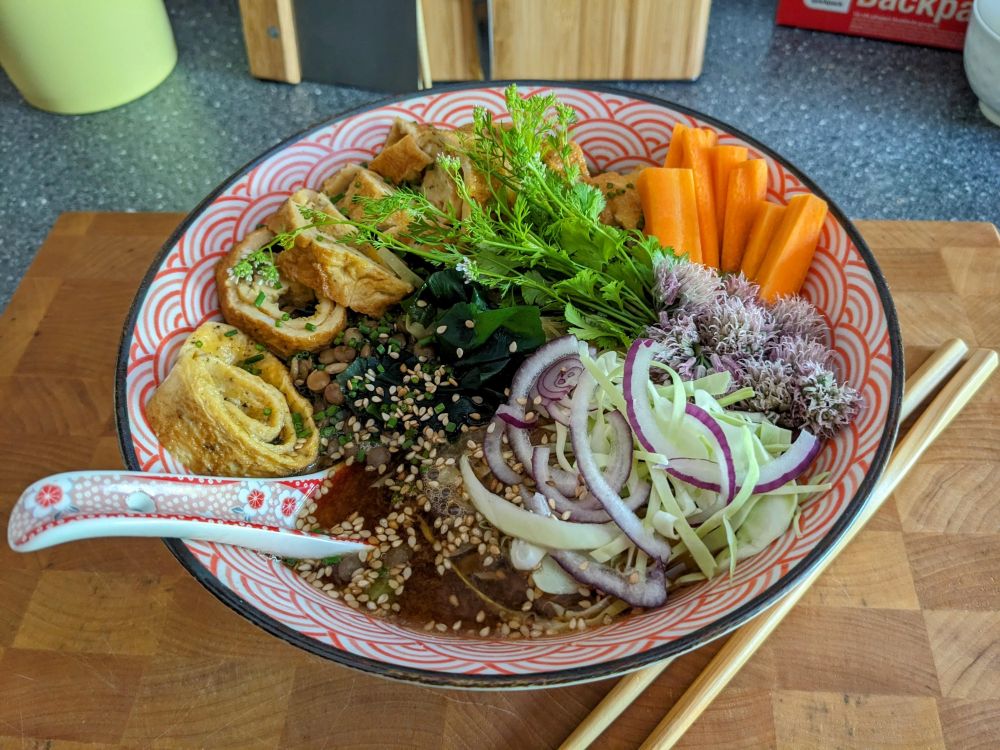 A noodle bowl filled with tamagoyaki, pardina lentils, miso soup, sweetheart cabbage, cilantro, carrots, chive flowers, wakame, roasted sesame seeds, and a homeopathic amount of thinly sliced red onions.