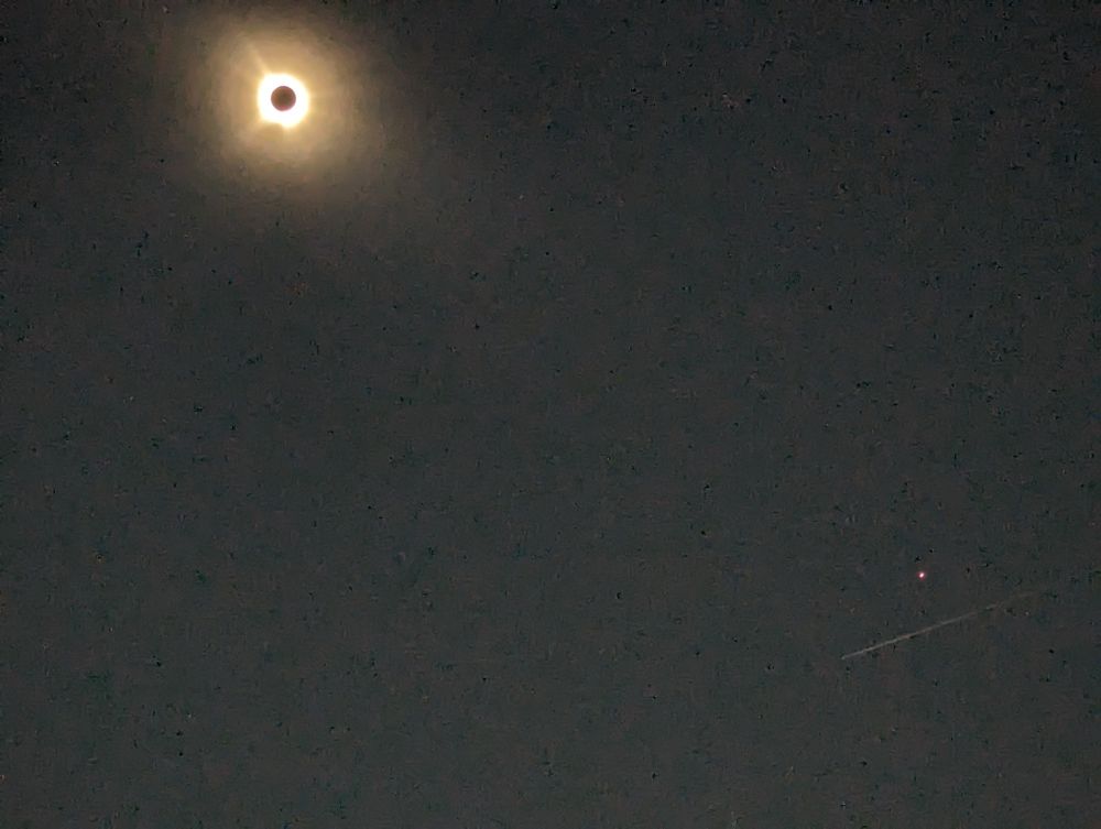 An eclipse picture of an irregular ring of white around a black circle in the upper left hand quadrant with a bright pinkish dot diagonally left and below the eclipse with a faint white line below the bright dot in the lower right corner.