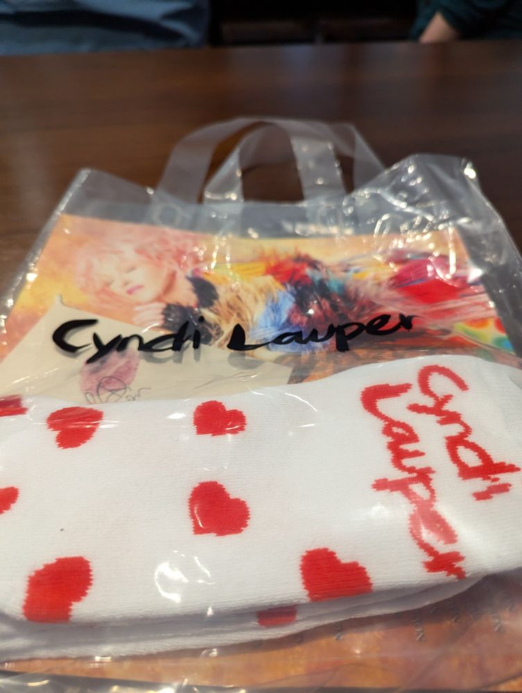 The swag bag for the Cyndi Lauper concert. It includes a white pair of socks with Cyndi Lauper's name and small poster of her in a very colorful outfit.