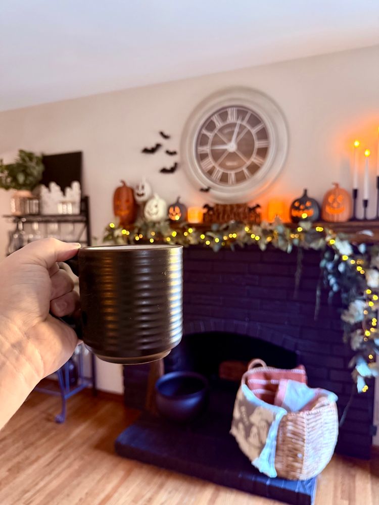 Holding up a cup of coffee, which is in focus. Out of focus is my Halloween mantle and winerack. 