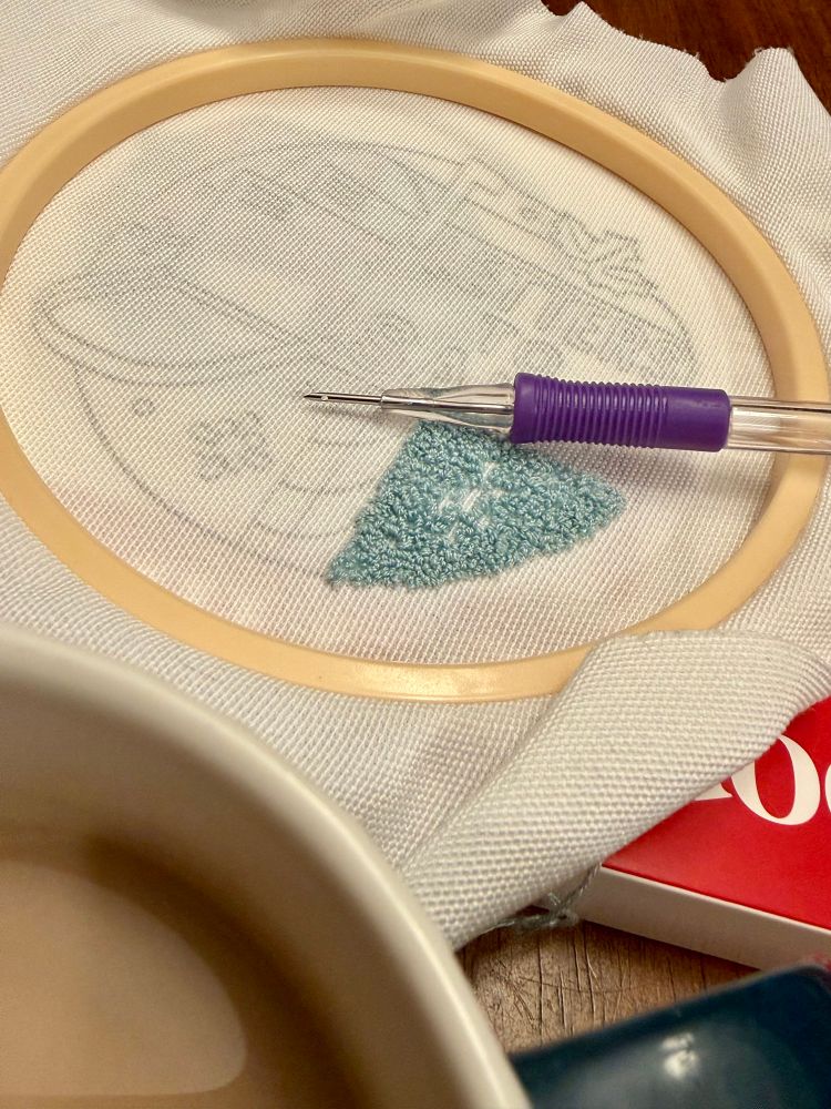 An embroidery hoop with a piece of fabric inside that has a design on it. A small section has been punch needled. The punch needle is resting on the embroidery hoop. 
