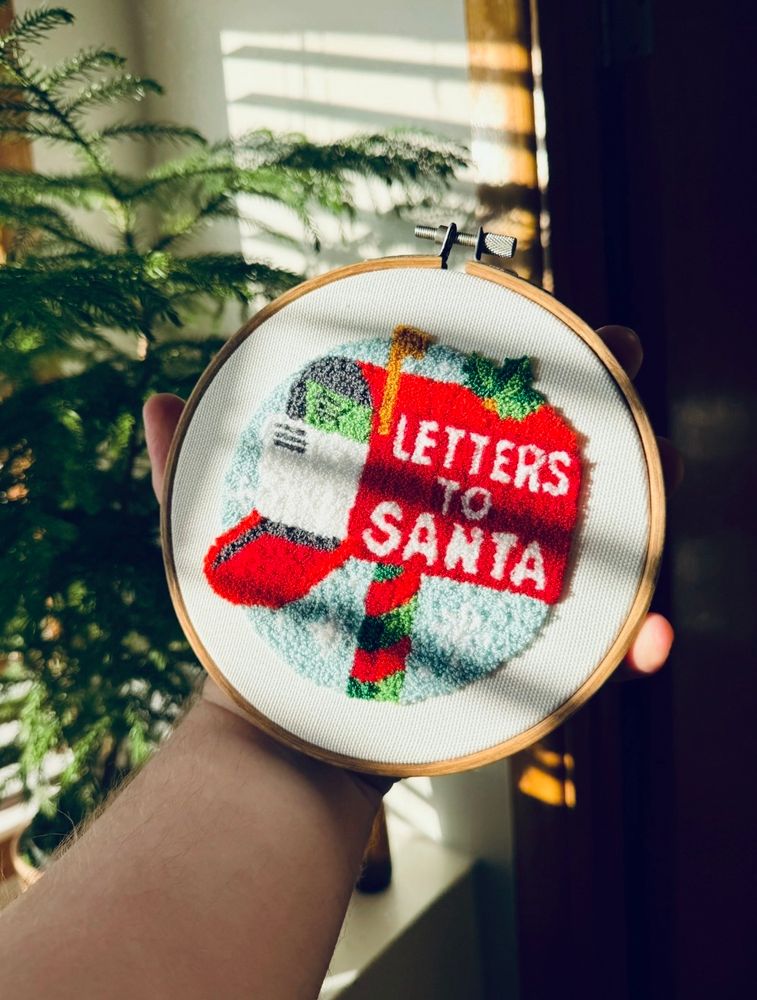 A Christmas themed punch needle in an embroidery hoop. It’s a red mailbox with a few parcels in it. Mailbox reads “Letters to Santa”. 