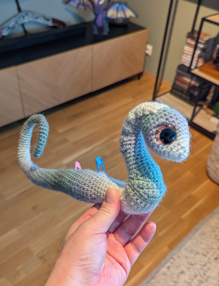 A hand holds a small, green striped, crochet dragon body. It has a head, but no other limbs or decorations.