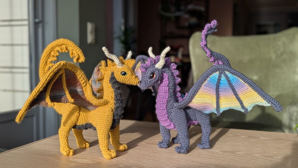 Two crocheted dragons stand nose to nose. One is yellow with grey and brown details, the other is blue with purple and pale blue and yellow details. 