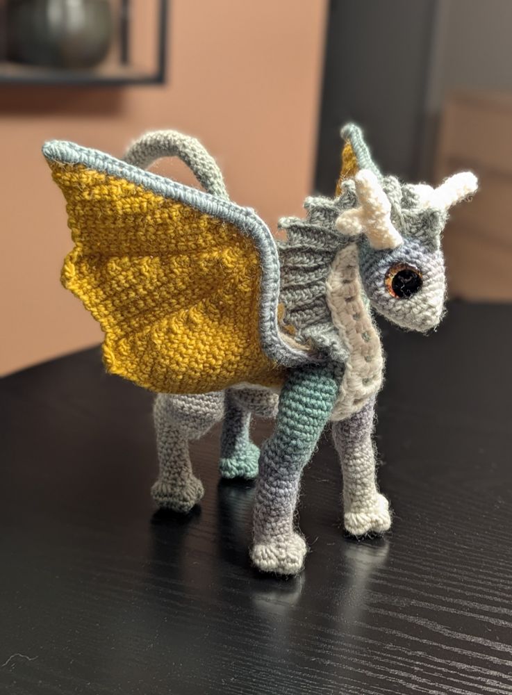 A crochet dragon. The body is green striped, the wings yellow, belly and horns are white. It has green frills on the neck, and large golden eyes, and is very cute.